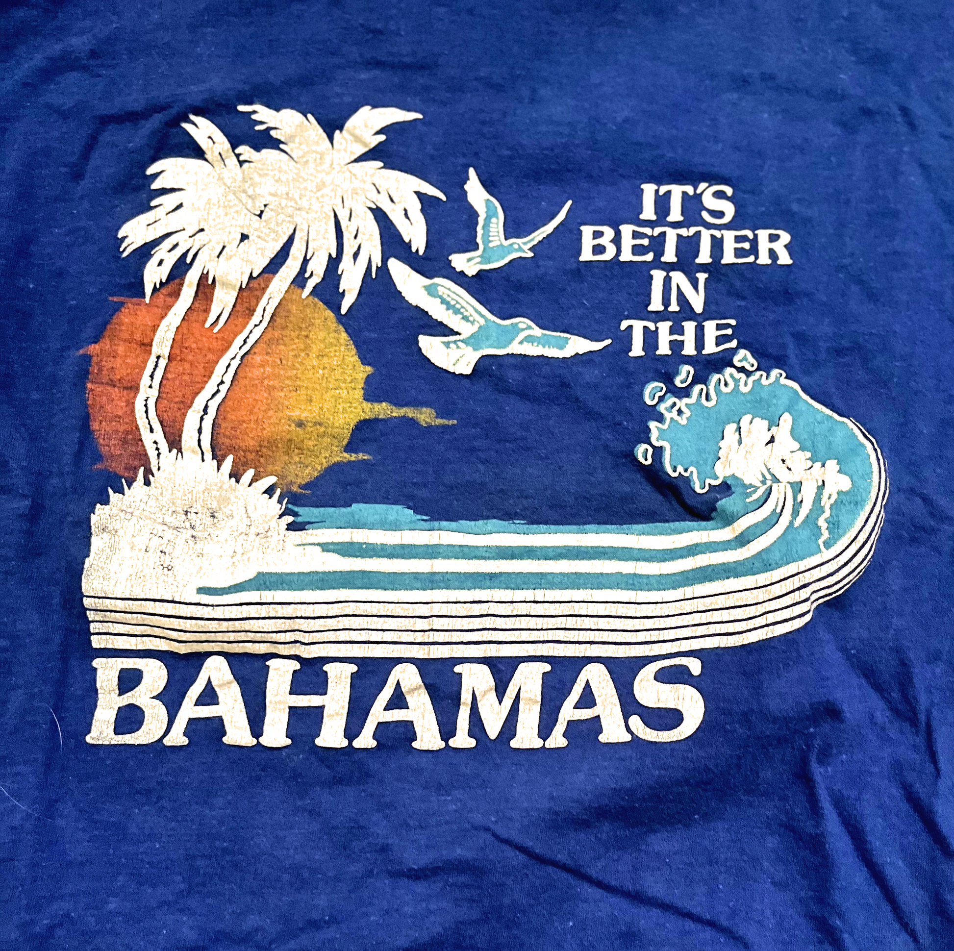 Back of blue t-shirt with 'its better in the Bahamas' graphic on white background.