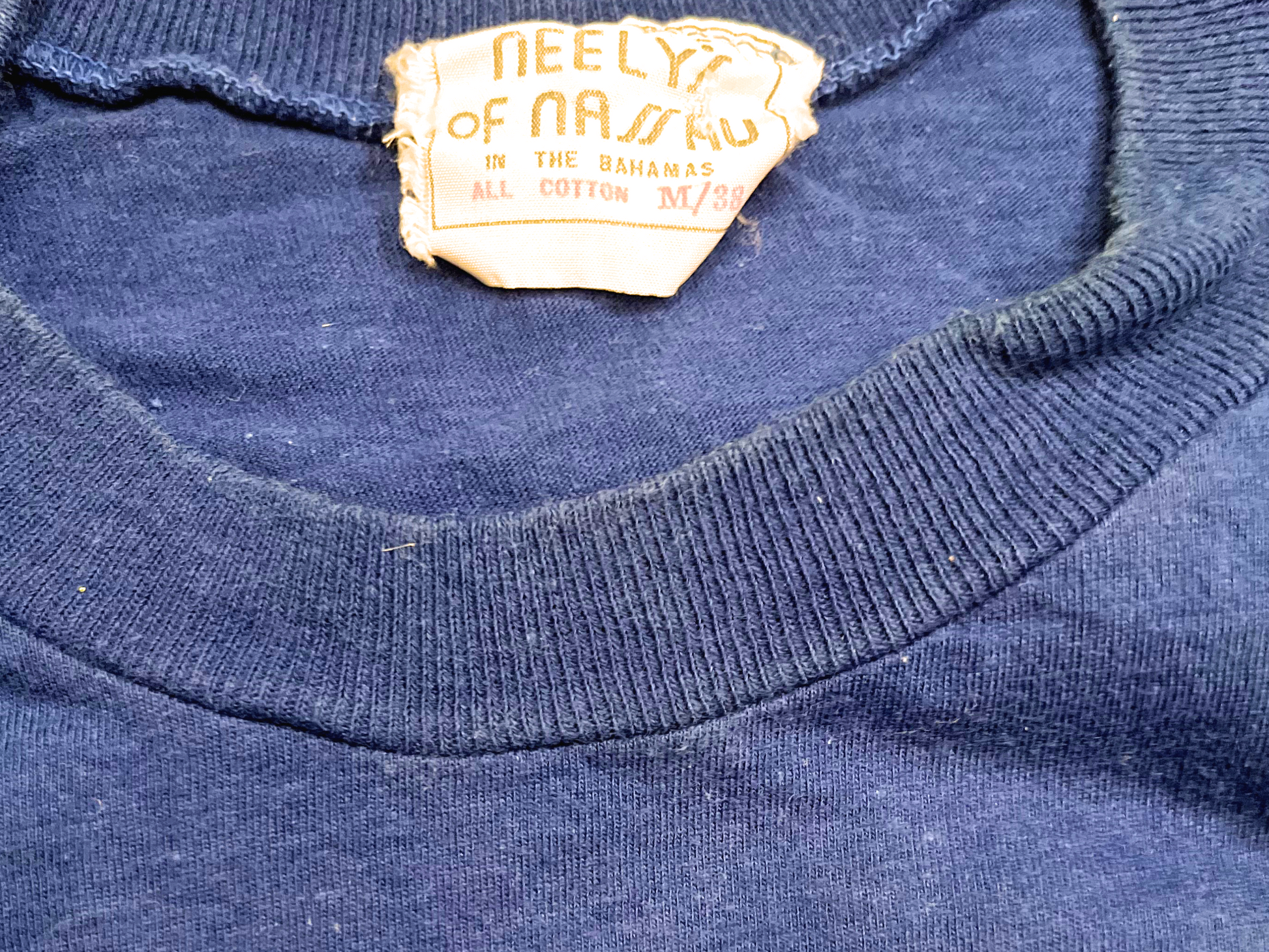 Neck label of blue t-shirt with 'its better in the Bahamas' graphic on white background.